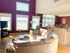 A living room with a beige sofa, a television, and a view of the beach through large windows.