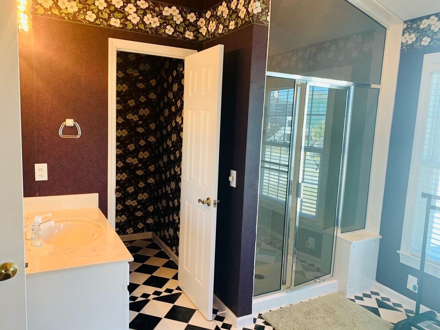 A bathroom with a white sink, a black and white checkered floor, a shower with a glass door, and a door leading to a dark room with floral wallpaper.