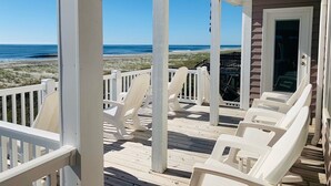 A covered deck with white chairs, a view of the beach, and a building with a glass door. - License To Chill 4 Bedroom Home by RedAwning (Supply)