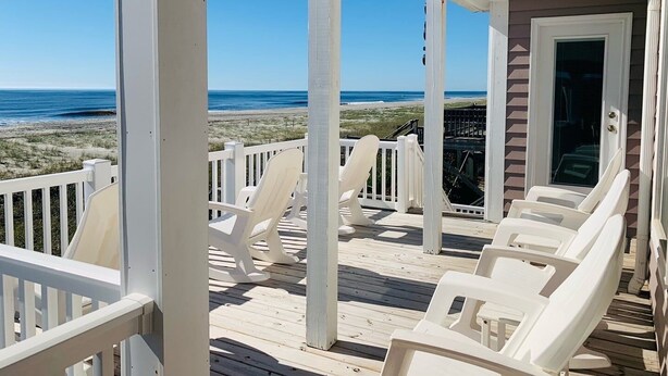 A covered deck with white chairs, a view of the beach, and a building with a glass door. - License To Chill 4 Bedroom Home by RedAwning (Supply)