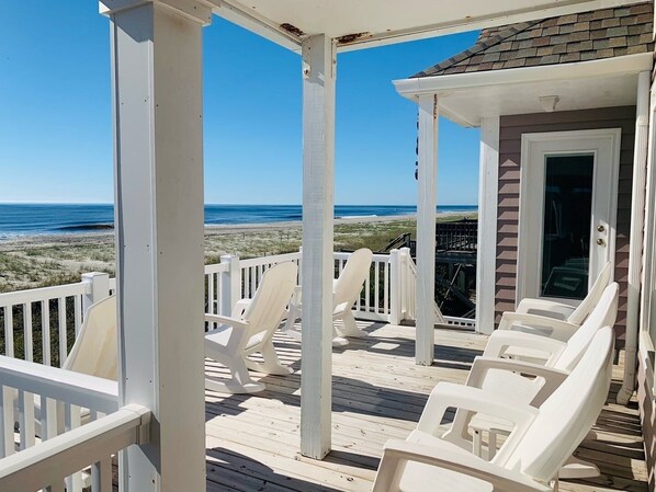 A covered deck with white chairs, a view of the beach, and a building with a glass door.
