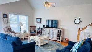 Un salon avec deux canapés bleus, une table basse blanche, un téléviseur posé sur un meuble blanc et un ventilateur de plafond.