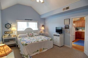 A bedroom with a bed, a television, a window, a clock, and a door leading to a kitchen. - Mclaurin Manna At Holden Beach 4 Bedroom Home by RedAwning (Supply)