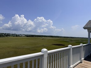 A view from a covered deck with a white railing, overlooking a grassy field and a distant line of houses under a partly cloudy sky.