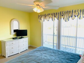A bedroom with a bed, a dresser, a television, a ceiling fan, and a sliding glass door to a balcony.