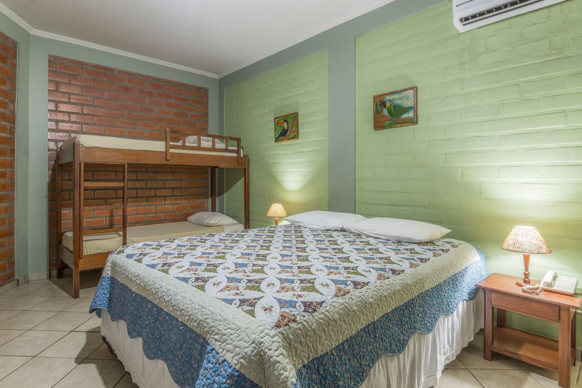 A room with a bunk bed, a quilted bedspread, a nightstand with a lamp, and two paintings on the wall.