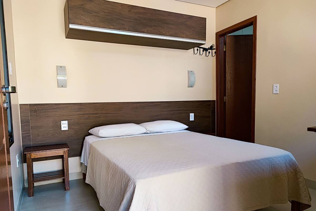 A neatly made bed with a wooden headboard, a small wooden stool, and a wall-mounted air conditioning unit.