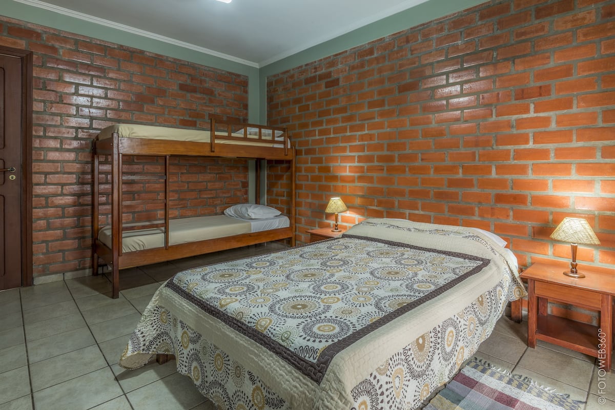 A room with a bunk bed, a single bed, a patterned bedspread, a small table with a lamp, and a brick wall.