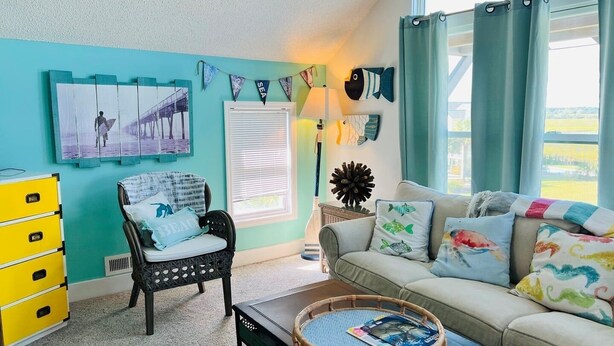 A living room with a teal wall, a black chair, a beige sofa, a yellow dresser, and a window with curtains. - Raising The Bar 5 Bedroom Home by RedAwning (Supply)