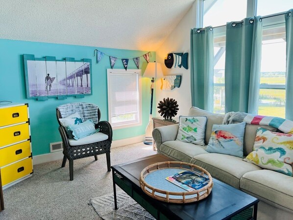 A living room with a teal wall, a black chair, a beige sofa, a yellow dresser, and a window with curtains. - Raising The Bar 5 Bedroom Home by RedAwning (Supply)