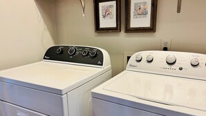 Two front-loading washers with control panels, a wooden shelf above, and two framed pictures on the wall. - Raising The Bar 5 Bedroom Home by RedAwning (Supply)