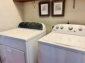 Two front-loading washers with control panels, a wooden shelf above, and two framed pictures on the wall. - Raising The Bar 5 Bedroom Home by RedAwning (Supply)