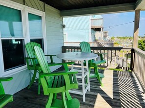A covered porch with a round table and four green chairs, overlooking a wooden deck and a residential area. - Raising The Bar 5 Bedroom Home by RedAwning (Supply)