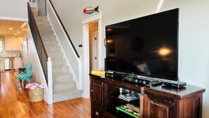 A living room with a wooden floor, a staircase, a flat-screen TV on a wooden stand, and a kitchen in the background. - Raising The Bar 5 Bedroom Home by RedAwning (Supply)