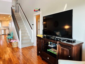A living room with a wooden floor, a staircase, a flat-screen TV on a wooden stand, and a kitchen in the background. - Raising The Bar 5 Bedroom Home by RedAwning (Supply)