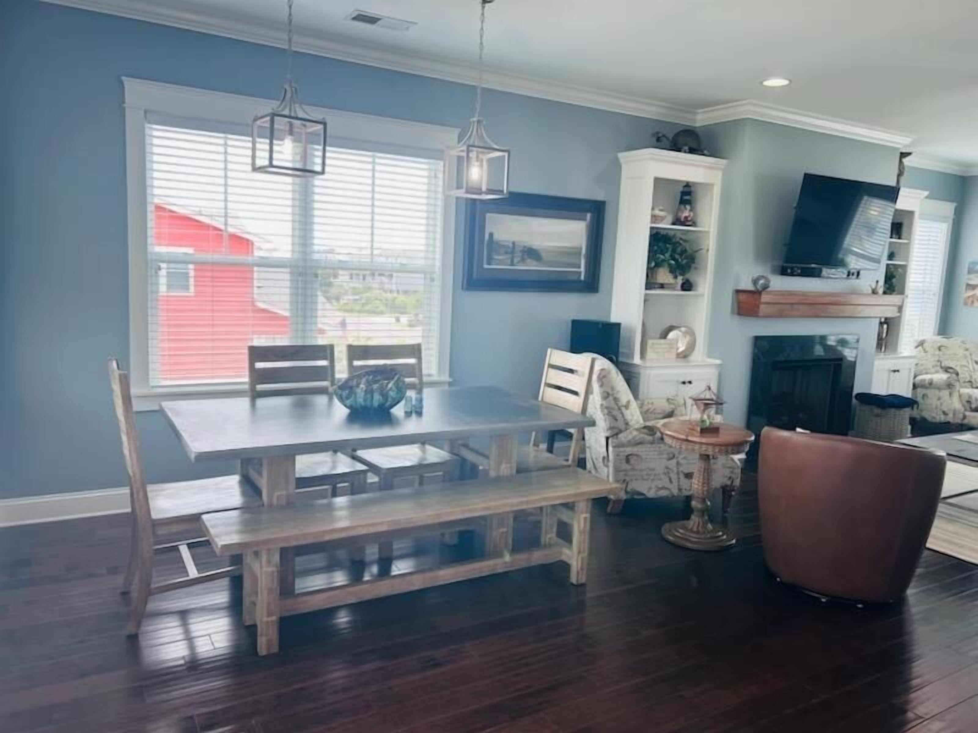 A dining area with a wooden table and chairs, a bench, pendant lights, and a view through a window with blinds.