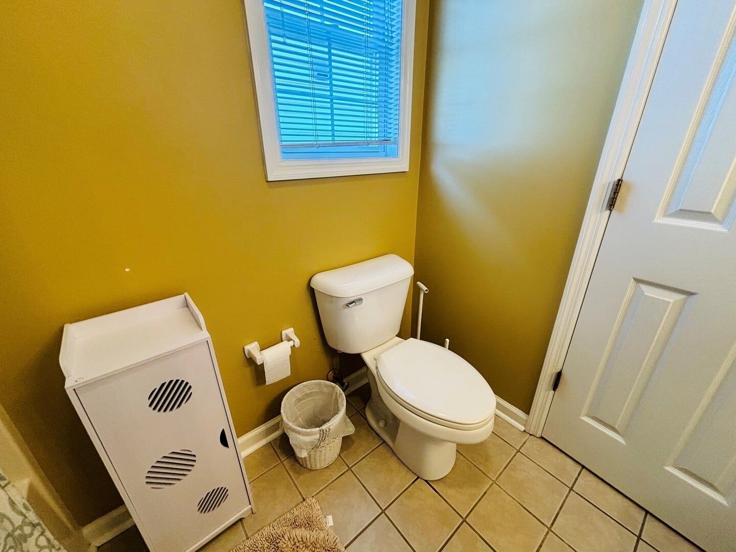 A bathroom with a toilet, a trash can, a window with blinds, and a door.