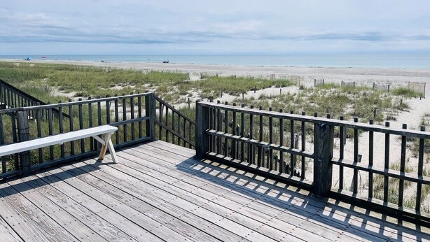 Una terraza de madera con un banco, con vista a una playa con dunas y una valla.