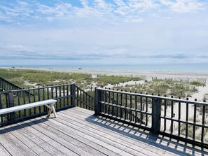 Una terraza de madera con un banco, con vista a una playa con dunas y una valla.