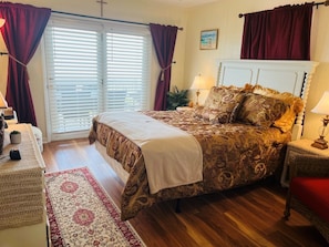 A bedroom with a bed, a nightstand, a lamp, a window with curtains, and a wall-mounted cross.