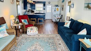 Una sala de estar acogedora con un sofá azul, un sillón rojo, una silla de mimbre, un otoman pequeño y redondo, y una alfombra estampada.