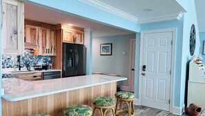 A kitchen with wooden cabinets, a black refrigerator, and a white countertop. There are three wicker stools with floral cushions. - Serenity's Star 4 Bedroom Home by RedAwning (Supply)