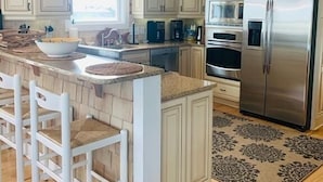 A kitchen with white cabinets, a stainless steel refrigerator, a microwave, and a built-in oven. There are two bar stools and a large island with a bowl. - Shore Beats Workin' 5 Bedroom Home by RedAwning (Supply)