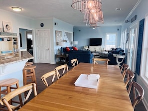 A dining area with a wooden table and chairs, a kitchen in the background, and a living room with a blue sofa. - Sunny Times 4 Bedroom Home by RedAwning (Supply)
