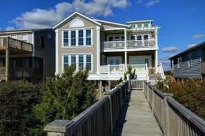 Ein mehrstöckiges Anwesen direkt am Strand mit einer Holzterrasse, die zum Eingang führt, einem Balkon im oberen Stockwerk und ungehindertem Blick auf den Ozean.