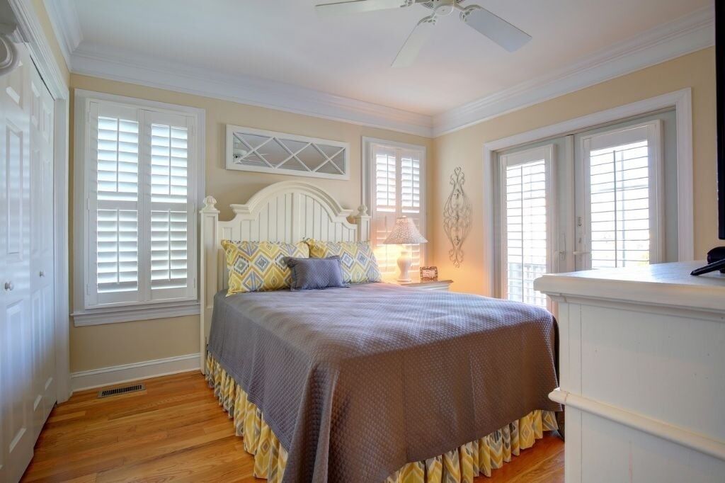 A bedroom with a bed, two windows with shutters, a ceiling fan, and a nightstand with a lamp.