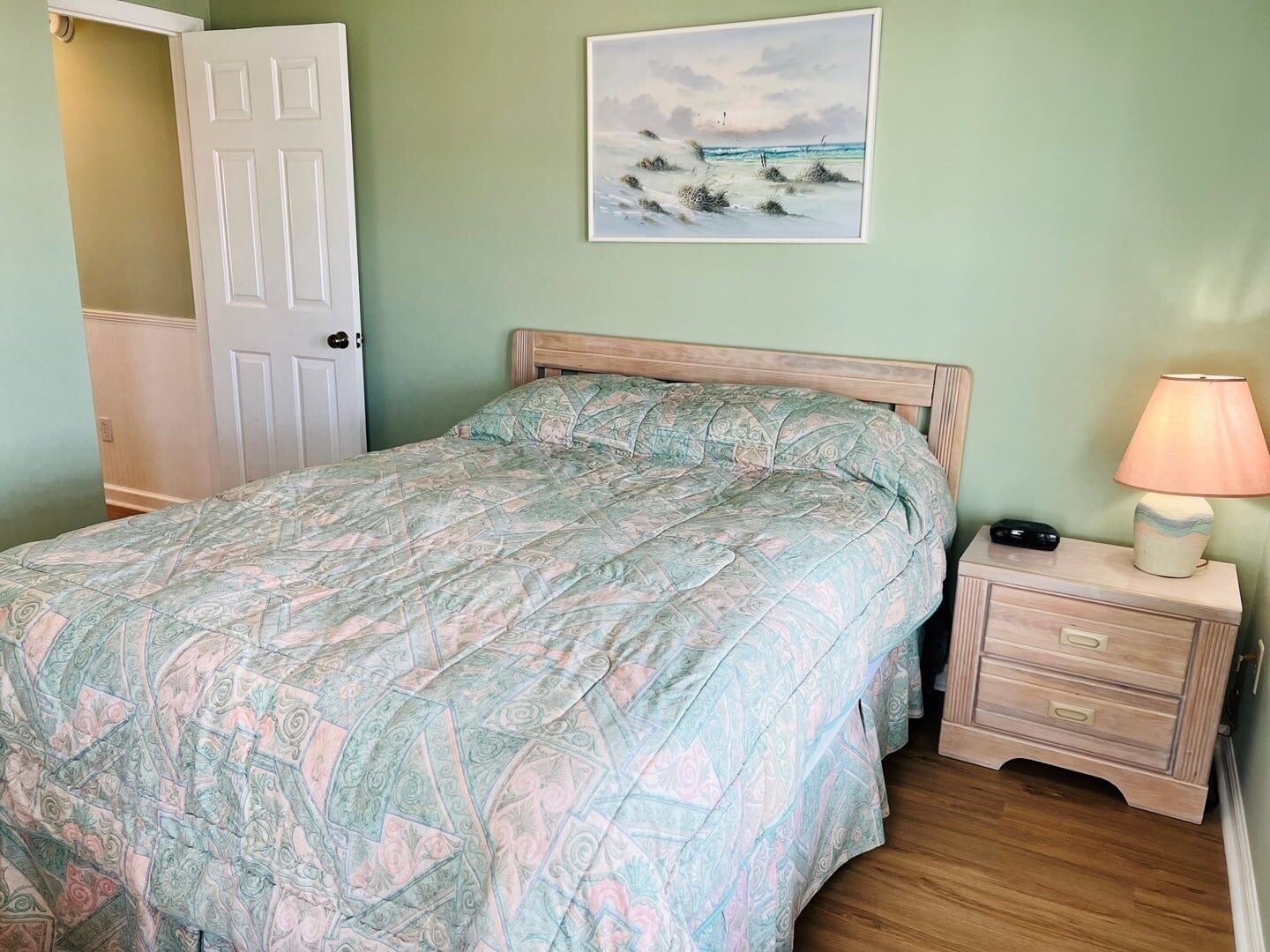 A bedroom with a bed, a nightstand with a lamp, a door, and a painting on the wall.