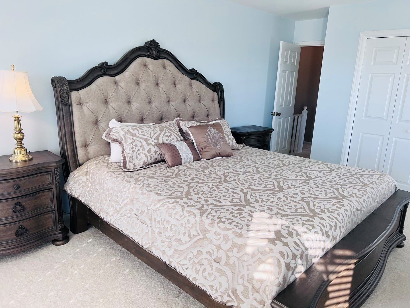 Una camera da letto con testiera trapuntata, copriletto decorato, abat-jour, cassettiera e una porta.