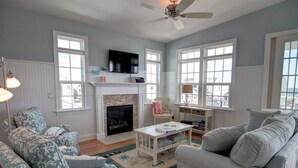 A living room with a fireplace, a ceiling fan, and large windows. - Welcome  4 Bedroom Home by RedAwning (Supply)