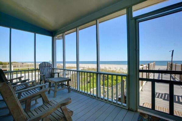A covered deck with wooden chairs and a table, overlooking a beach and boardwalk.