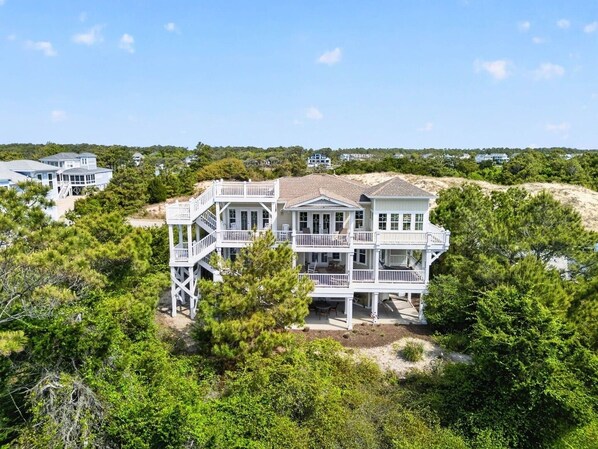 A large, multi-story beachfront property with a prominent balcony area, surrounded by dense greenery.