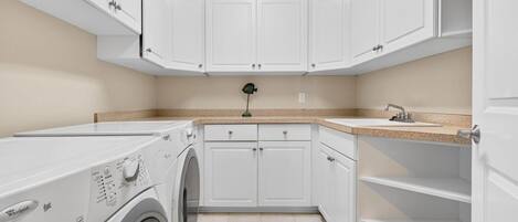 A laundry room with a front-loading washer and dryer, white cabinetry, a countertop with a sink, and a small green decorative item on the wall.