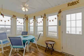A room with a table and chairs, a small stool, a ceiling fan, and a view of the beach through large windows.