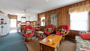 Ett mysigt rum med ett träpanelat tak, en takfläkt, ett matbord, flätade stolar, en soffa och ett kök i bakgrunden.