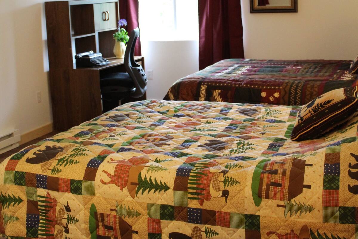 A bedroom with a large bed covered in a colorful quilt, a desk with a typewriter, a vase with flowers, and a window with curtains.
