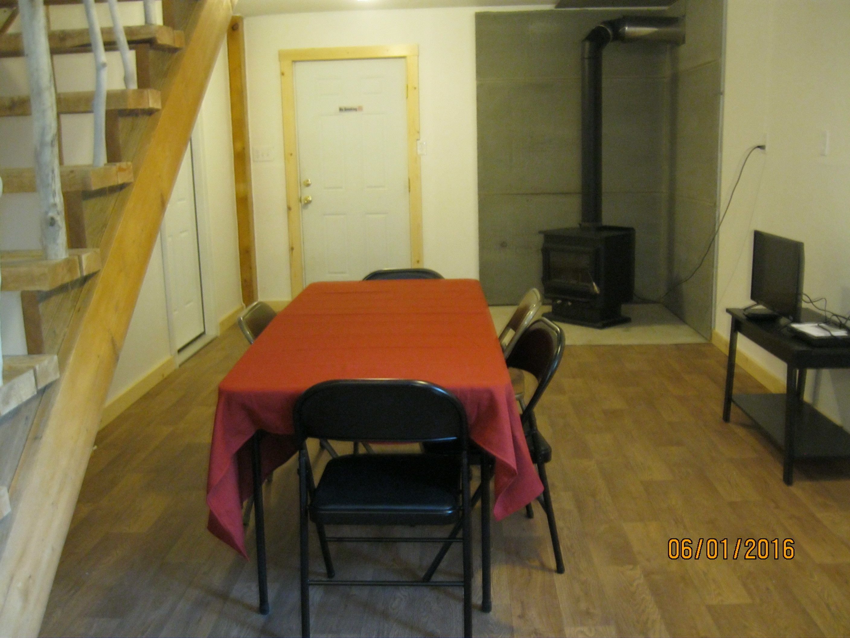 Una habitación con una escalera de madera, una mesa de comedor con sillas, un televisor sobre un escritorio y un estufa de leña.