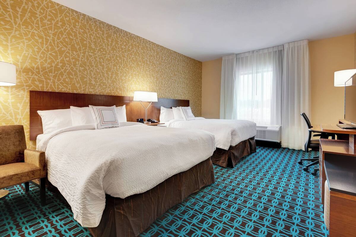 A hotel room with two beds, a desk, a chair, and a large window with curtains.