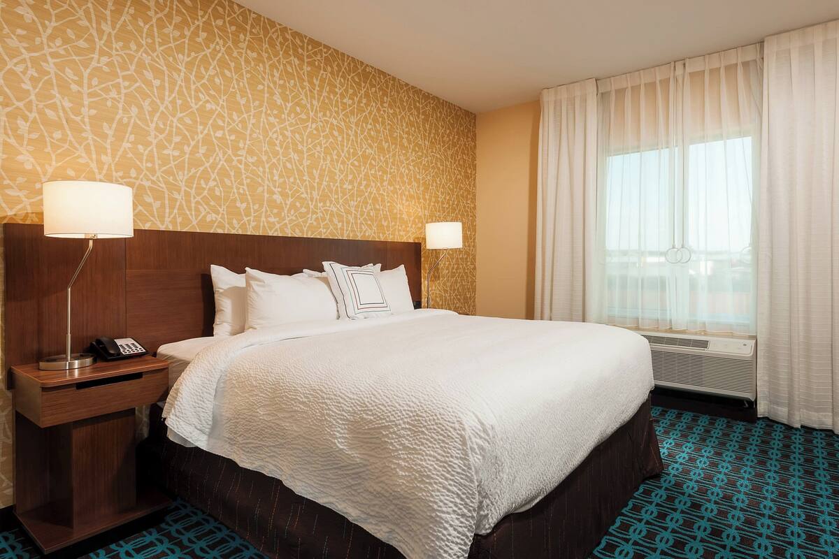A hotel room with a large bed, two bedside lamps, a nightstand with a phone, a window with curtains, and a patterned wall.