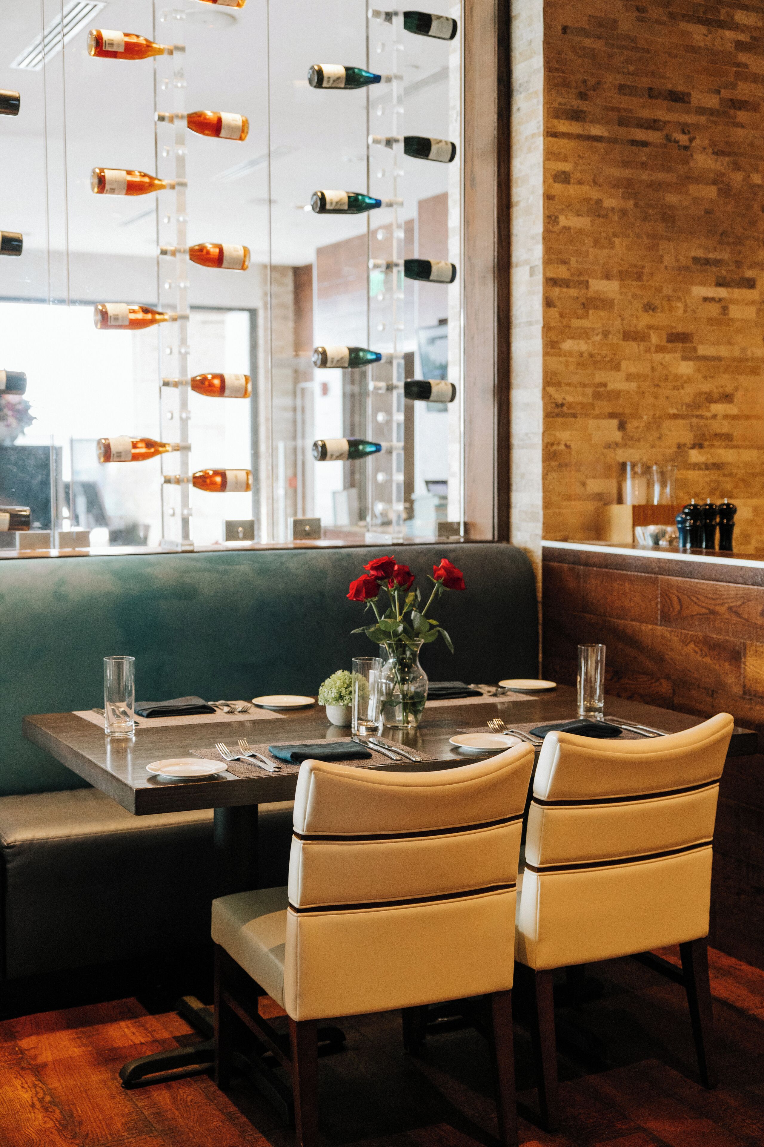 A restaurant interior with a brick wall, a glass display of bottles, a table set for two with chairs, and a vase with red roses.