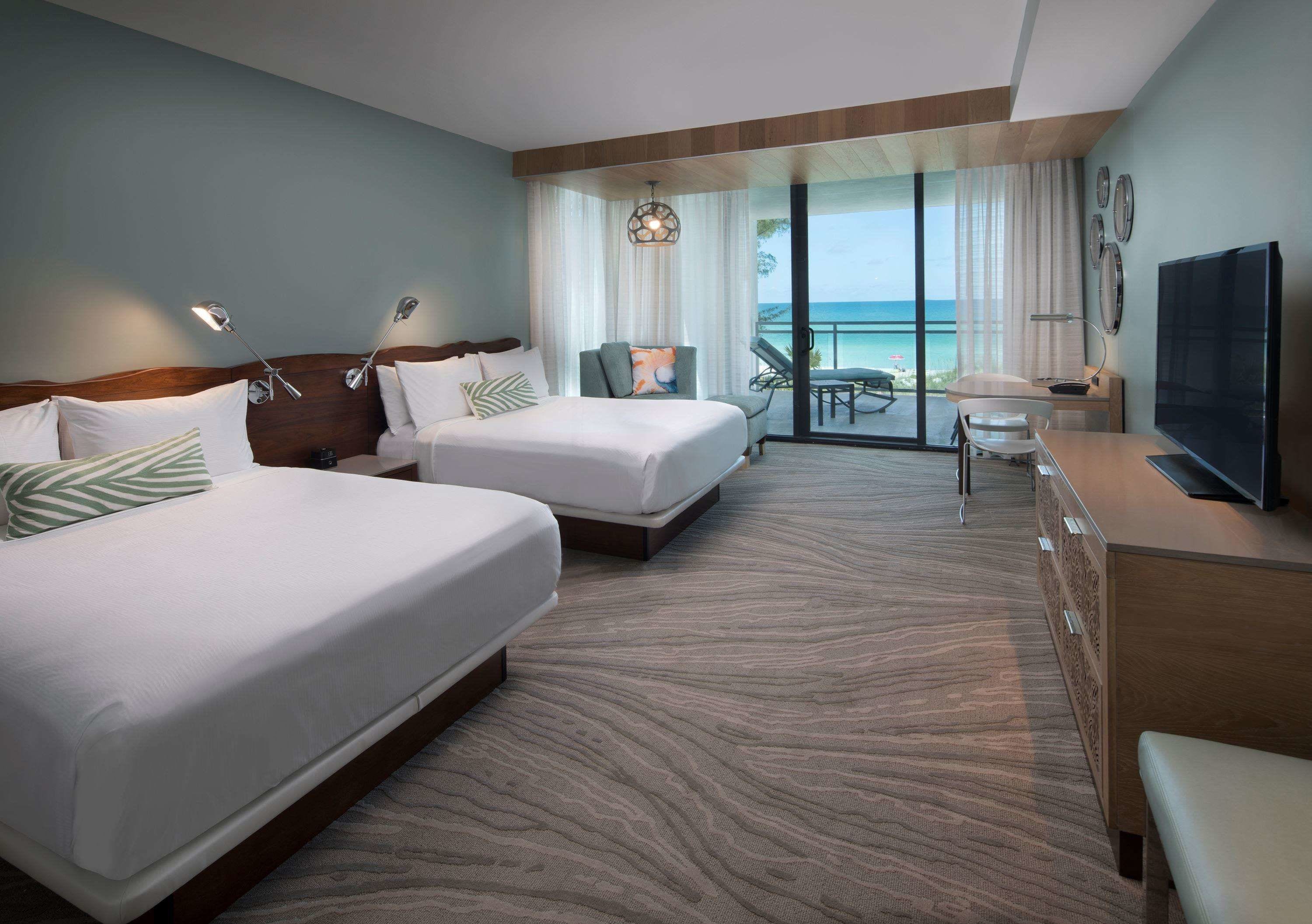 A hotel room with two beds, a TV, a desk, and a balcony with a view of the ocean.