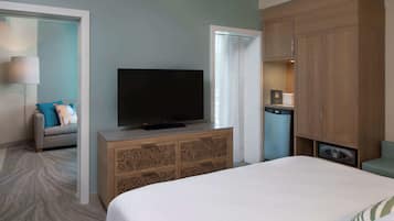 Habitación de hotel con cama, televisor sobre un mueble de madera, sofá con cojines y una mesita con lámpara.