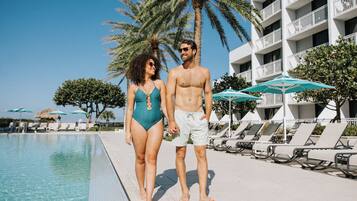 A couple walking by a poolside with lounge chairs and umbrellas, palm trees, and a building with balconies.