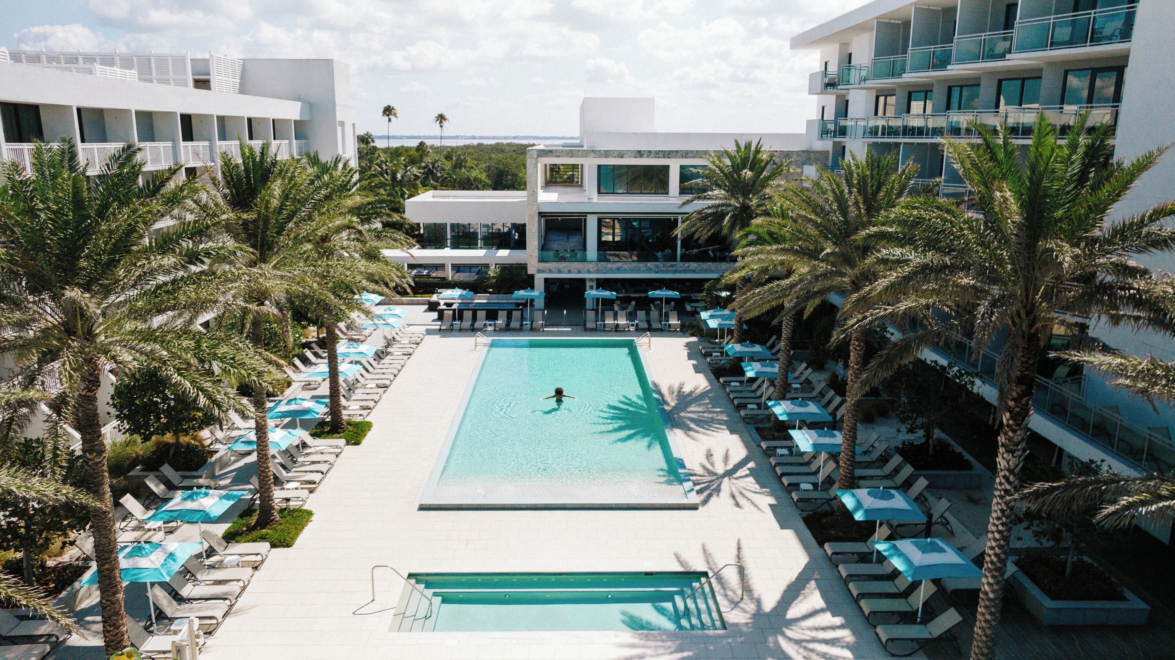 A pool area with sun loungers and umbrellas, surrounded by palm trees and modern buildings.