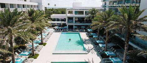 A pool area with sun loungers and umbrellas, surrounded by palm trees and modern buildings.