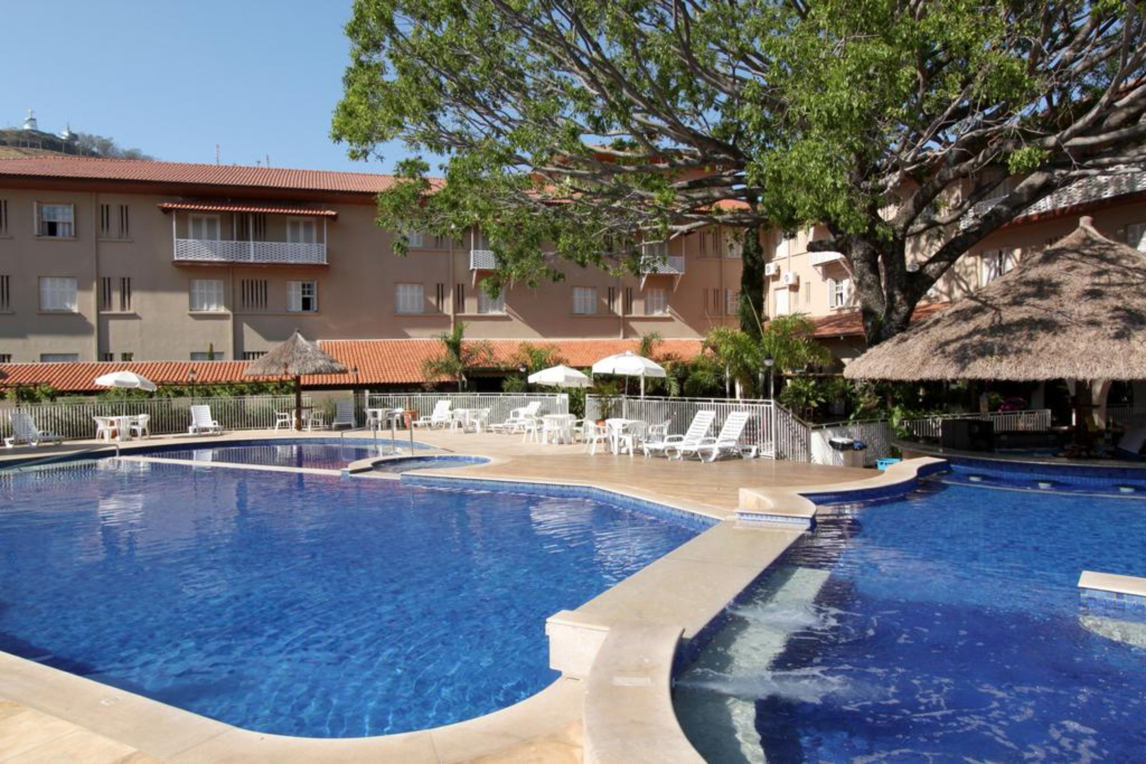 A swimming pool with lounge chairs and umbrellas, surrounded by buildings with balconies and a tree.