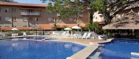 A swimming pool with lounge chairs and umbrellas, surrounded by buildings with balconies and a tree.